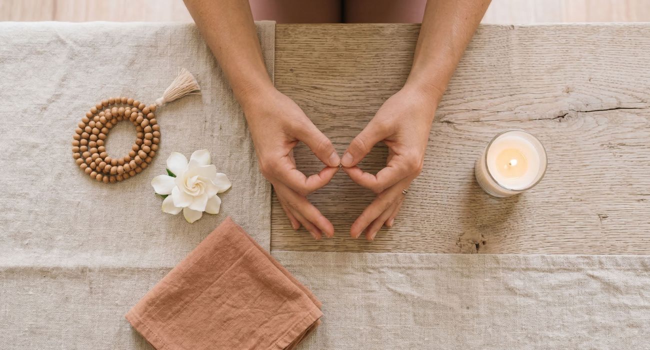 Hände einer Frau in Meditationsmudra neben Mala-Perlen, einer weißen Blume und einer Kerze auf Leinen