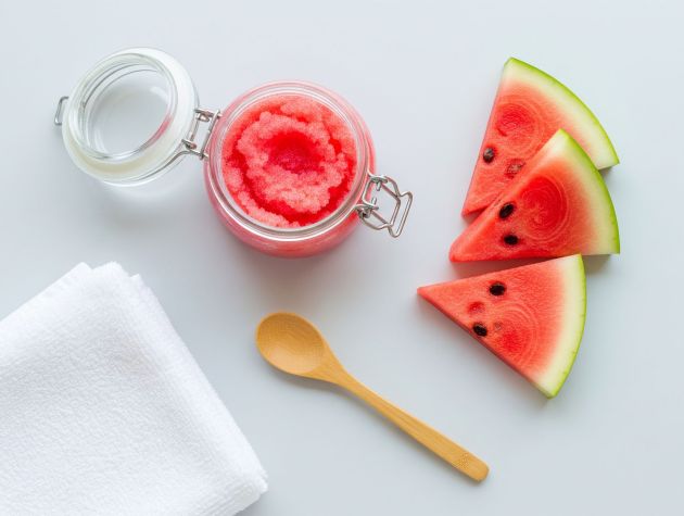 Tarro de exfoliante de azúcar y sandía con trozos de sandía y cuchara de madera sobre fondo claro.