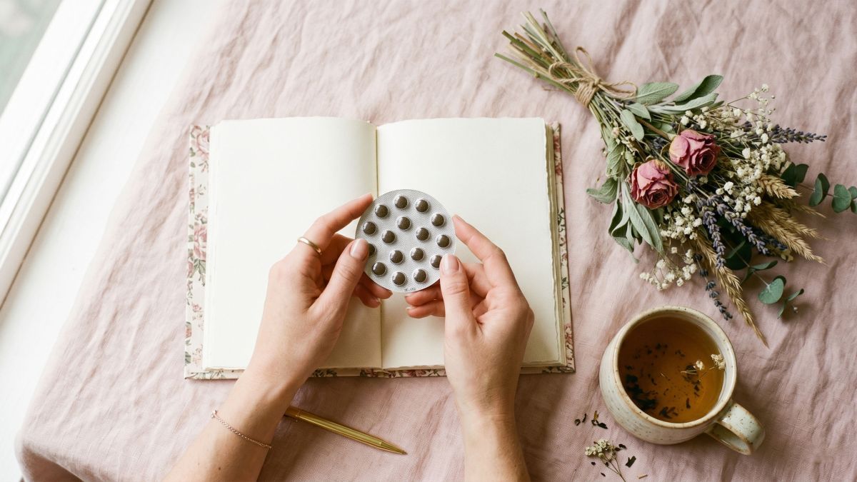 Manos de una mujer sosteniendo un paquete de píldoras anticonceptivas junto a un diario y té herbal sobre un fondo pastel