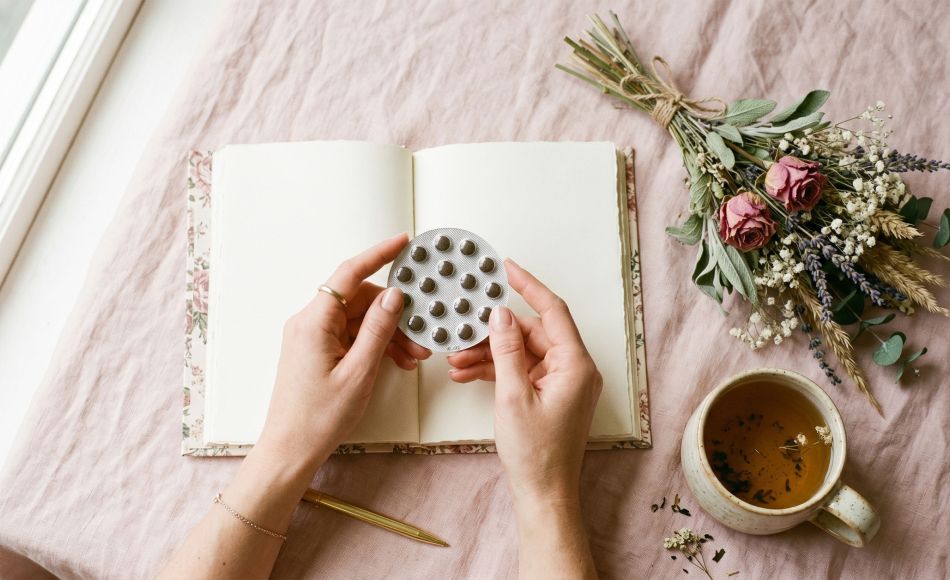 Manos de una mujer sosteniendo un paquete de píldoras anticonceptivas junto a un diario y té herbal sobre un fondo pastel