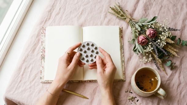 Manos de una mujer sosteniendo un paquete de píldoras anticonceptivas junto a un diario y té herbal sobre un fondo pastel