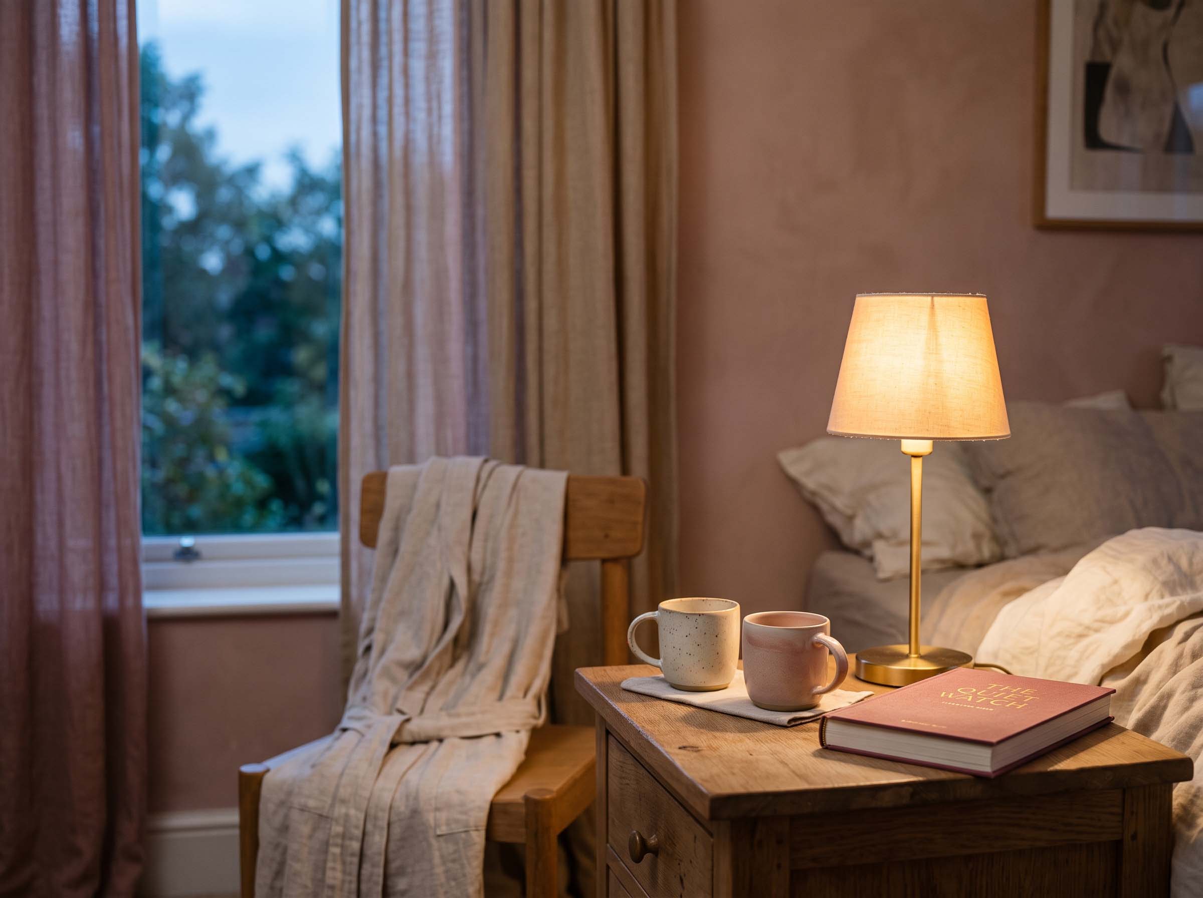 Bedside lamp glowing softly beside a folded robe and two mugs in a quiet evening bedroom