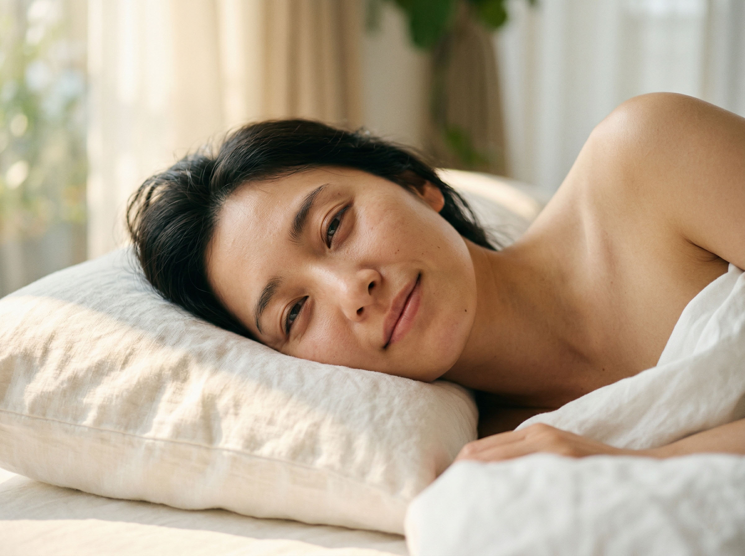 柔らかな朝の光の中で穏やかに目覚める女性、白いリネンのシーツ