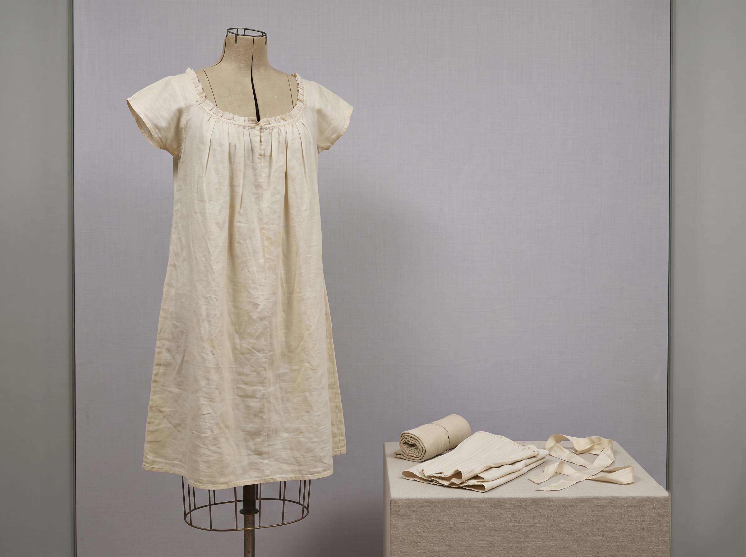 Antique white linen chemise displayed on a dress form beside folded historical undergarments, illustrating the history of womens nightwear