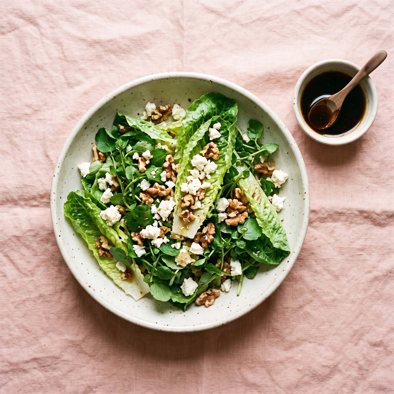 Fresh romaine and watercress salad with walnuts and crumbled cheese in a pale ceramic bowl on a soft pink surface