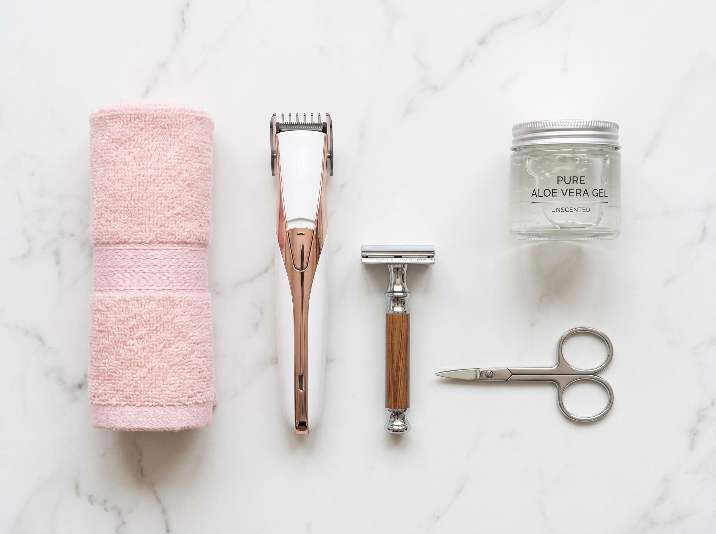 Flat lay of feminine grooming tools including a trimmer, soft towels, aloe gel, and a single-blade razor on a marble surface