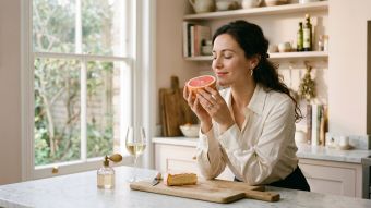 Woman breathing in the scent of a grapefruit at a sunlit kitchen counter beside wine and cheese, illustrating the chemistry of human scent