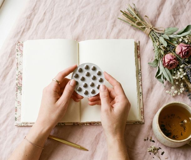 Manos de una mujer sosteniendo un paquete de píldoras anticonceptivas junto a un diario y té herbal sobre un fondo pastel