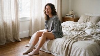 Woman sitting on the edge of an unmade bed in an oversized cotton t-shirt and mismatched socks in soft morning light, illustrating what women actually wear to sleep