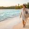 Una mujer camina por una prístina costa tropical sosteniendo un sombrero para el sol y una bolsa de playa tejida.