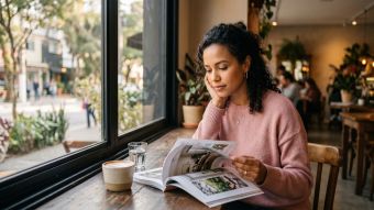 Mulher sentada pensativamente junto à janela de um café lendo uma revista, luz natural suave, expressão contemplativa e tons quentes