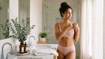 Mulher com um roupão de linho numa casa de banho iluminada com produtos suaves de cuidados da pele, representando cuidados íntimos tranquilos em casa.