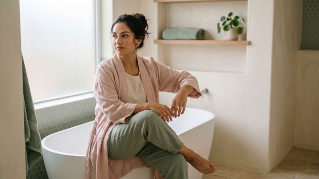 Mujer sentada en un baño iluminado por el sol, reflexionando — normalizando la conciencia corporal y las rutinas de higiene diaria