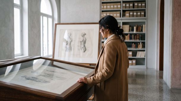 Mujer con abrigo color camel observando un grabado anatómico del siglo XIX enmarcado en un archivo museístico, que ilustra la historia de cómo se representaba la anatomía femenina