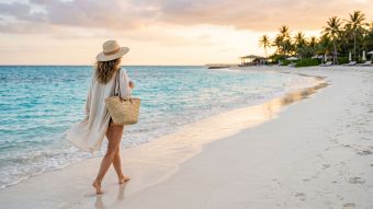 A woman walking along a pristine tropical shoreline holding a sun hat and a woven beach bag.