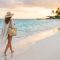 A woman walking along a pristine tropical shoreline holding a sun hat and a woven beach bag.