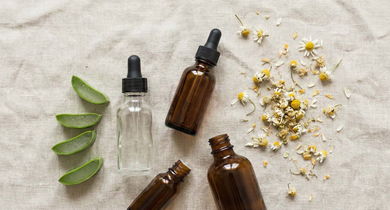 Eine Sammlung natürlicher Hautpflege-Inhaltsstoffe wie Aloe Vera, Kamille und botanische Öle auf einer hellrosa Leinenoberfläche.