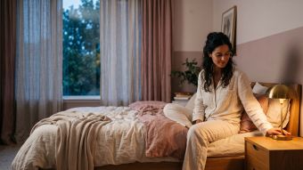 Mulher usando pijama creme acendendo uma luminária de cabeceira ao anoitecer, ilustrando os sinais não verbalizados que os casais usam para a intimidade.