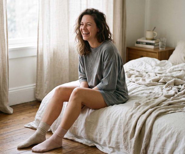 Woman sitting on the edge of an unmade bed in an oversized cotton t-shirt and mismatched socks in soft morning light, illustrating what women actually wear to sleep