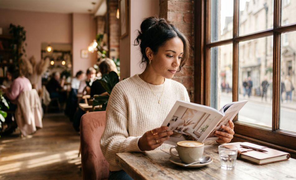 Woman sitting thoughtfully at a café window reading a magazine, soft natural light, contemplative expression, warm tones