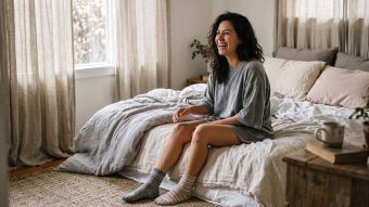 Mujer sentada en el borde de una cama sin hacer, con una camiseta de algodón extragrande y calcetines desparejados bajo una suave luz matutina, ilustrando lo que realmente usan las mujeres para dormir.