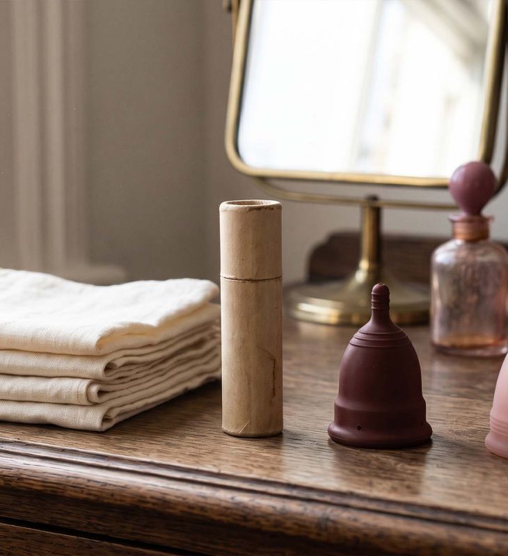 History of period products timeline shown on a sunlit dressing table with folded linen cloths, a vintage cardboard applicator, a rubber menstrual cup and a modern silicone cup