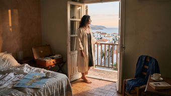 Woman in a linen robe at a hotel balcony doorway in morning light, reflecting on why travel doesn't restore closeness in a marriage