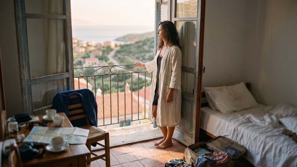 Mulher usando um roupão de linho na porta da varanda de um hotel à luz da manhã, refletindo sobre por que viajar não restaura a proximidade em um casamento