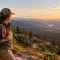 Woman hiker pausing beside a granite boulder on a mountain trail at golden hour with a rose bandana tied to her daypack