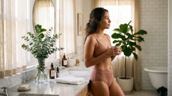 Mujer con una bata de lino en un baño luminoso junto a productos suaves para el cuidado de la piel, representando un cuidado íntimo tranquilo en casa.