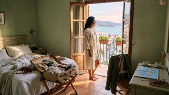 Mujer con bata de lino en la entrada del balcón de un hotel a la luz de la mañana, reflexionando sobre por qué viajar no restaura la cercanía en un matrimonio