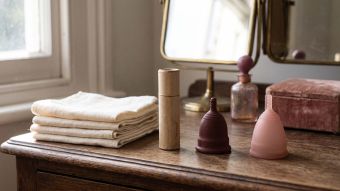 History of period products timeline shown on a sunlit dressing table with folded linen cloths, a vintage cardboard applicator, a rubber menstrual cup and a modern silicone cup