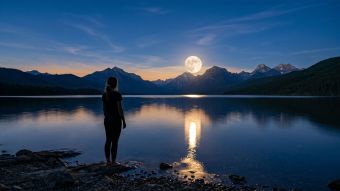 Full moon rising over calm ocean water at blue hour, with the silhouette of a woman standing at the shoreline looking up — symbolizing the ancient connection between the lunar cycle and the menstrual cycle